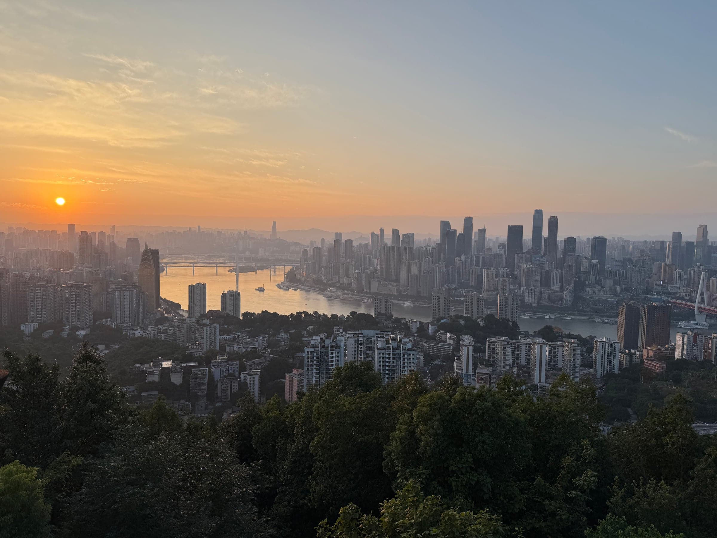 Coucher de soleil sur le Yangtsé et la skyline de Chongqing sous un ciel orangé, vue depuis les hauteurs de la rive sud