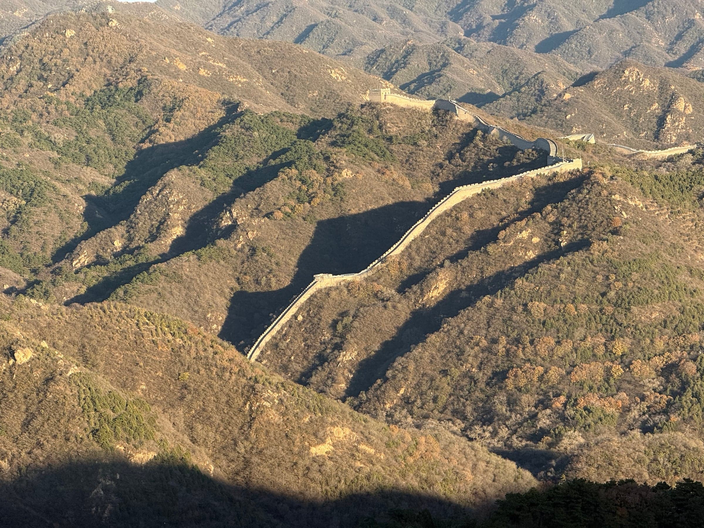 La Grande Muraille de Chine descendant un versant boisé aux couleurs d'automne dans la lumière dorée de fin de journée, crêtes montagneuses successives et tours de garde le long du rempart