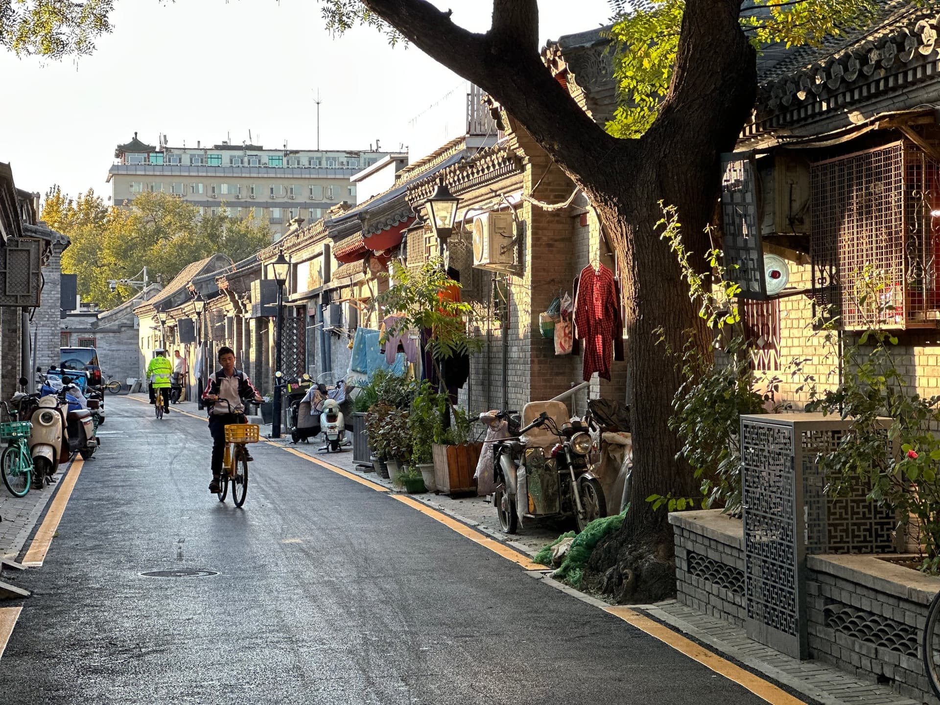 Ruelle traditionnelle des hutongs de Pékin tôt le matin, vélo au loin, lanternes rouges suspendues, vie de quartier qui se réveille