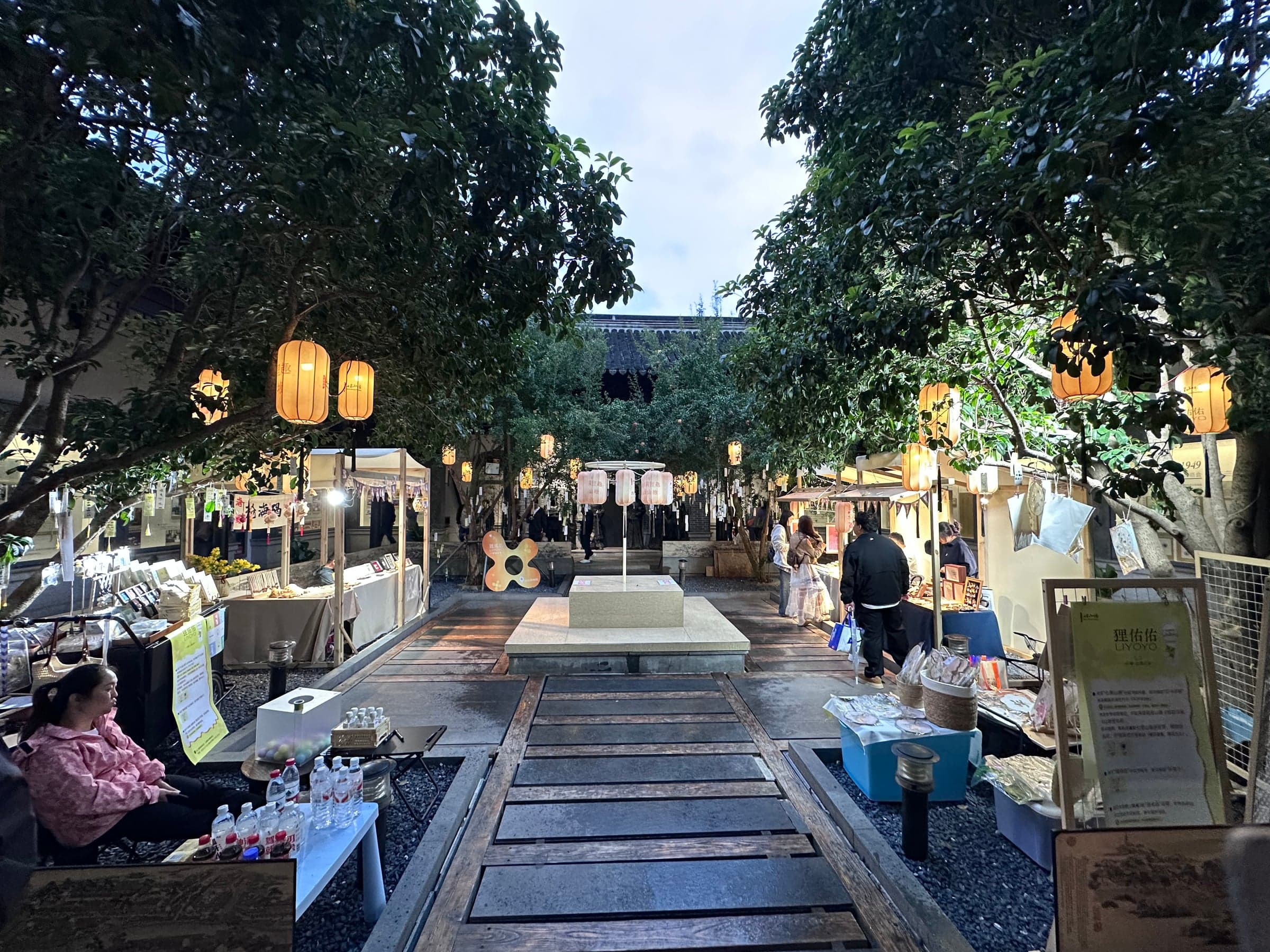 Marché nocturne dans une cour arborée de Suzhou, stands éclairés de lanternes, vendeurs derrière leurs étals